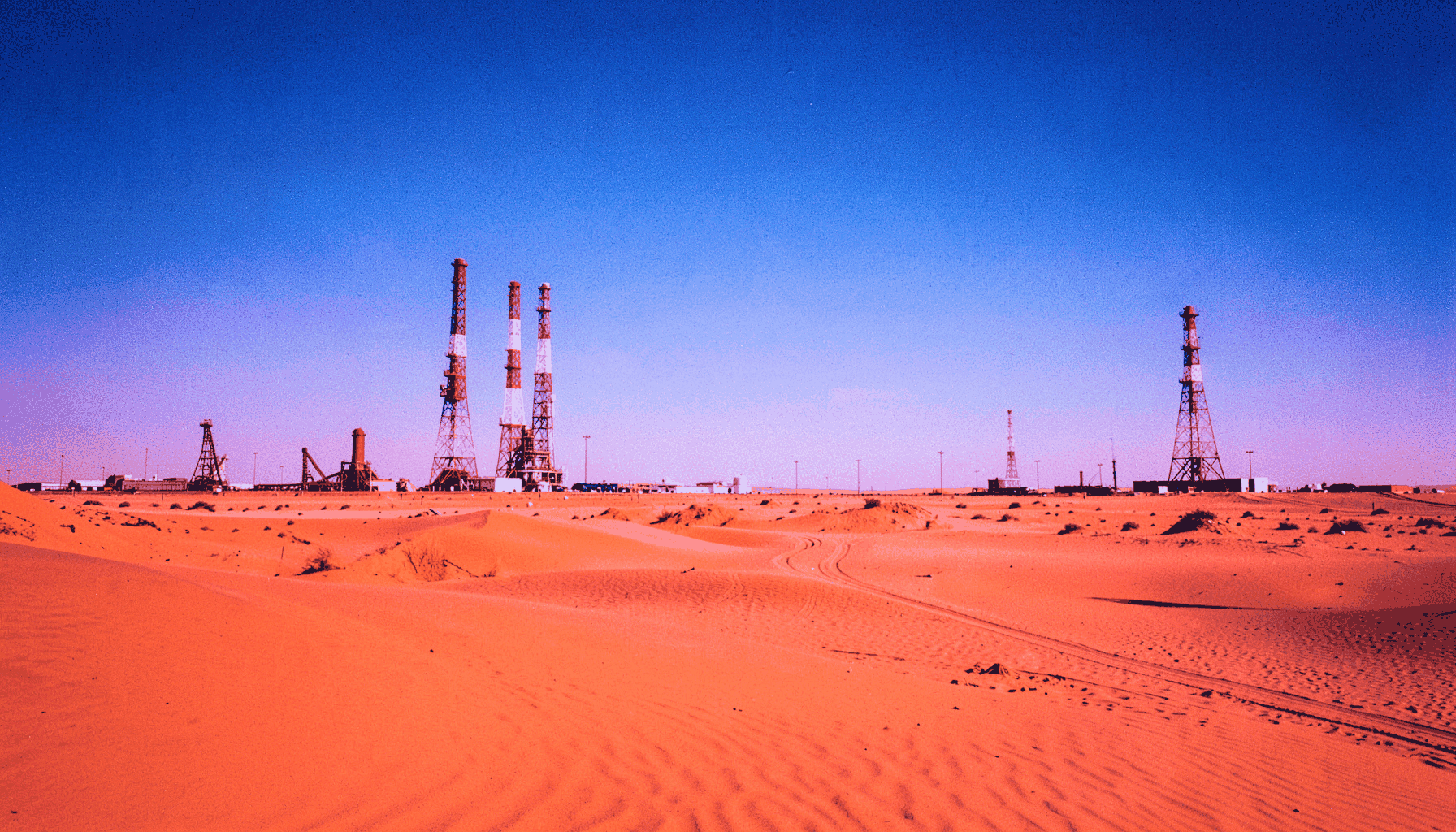 Paisagem desértica com torres de petróleo ao fundo, tons quentes de areia e céu azul intenso